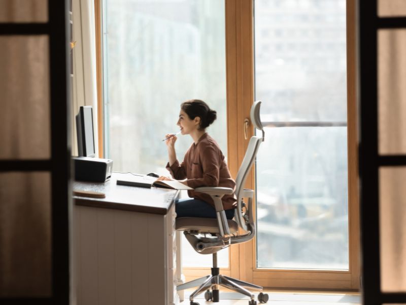 Žena sedi u ergonomskoj stolici pored velikog prozora i radi za računarom u dobro osvetljenom radnom prostoru.