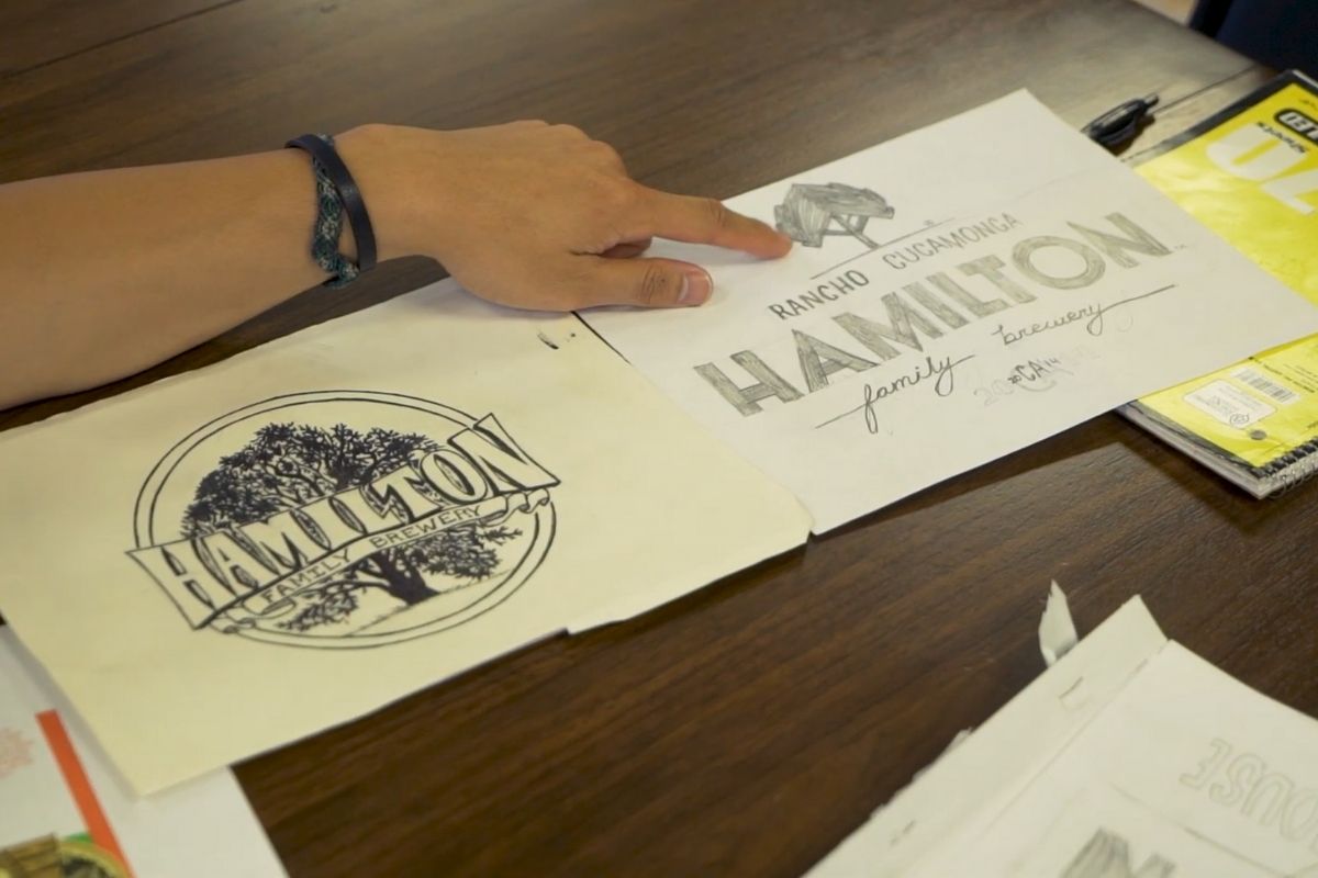 Ruka pokazuje na skicu logotipa „Hamilton Family Brewery“ na stolu