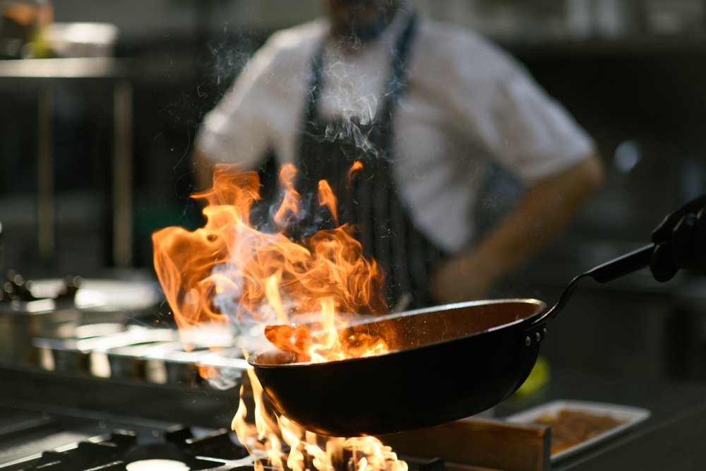 Kuvar priprema hranu u profesionalnoj kuhinji dok plamen zahvata tiganj