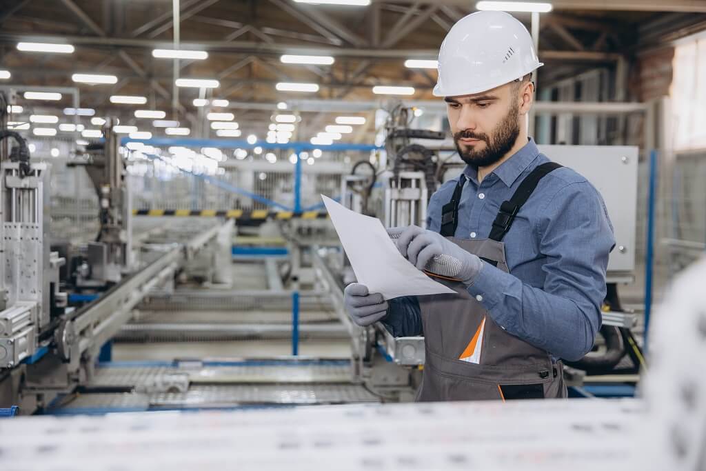 Radnik u proizvodnom pogonu sa zaštitnom kacigom i rukavicama pregleda dokumentaciju u industrijskom okruženju